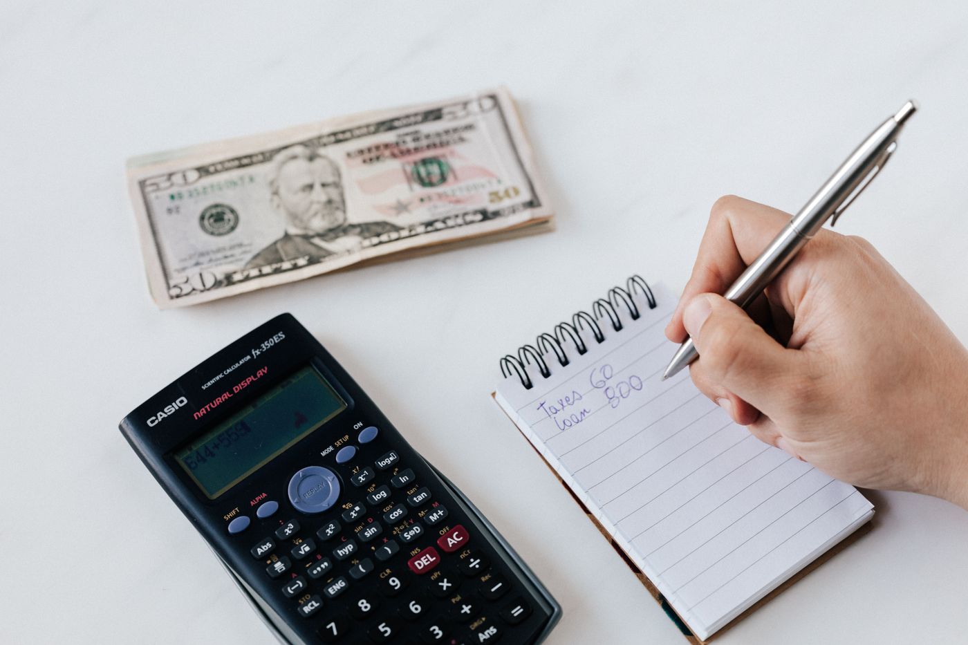 A hand writing in a notebook next to a calculator on a white desk