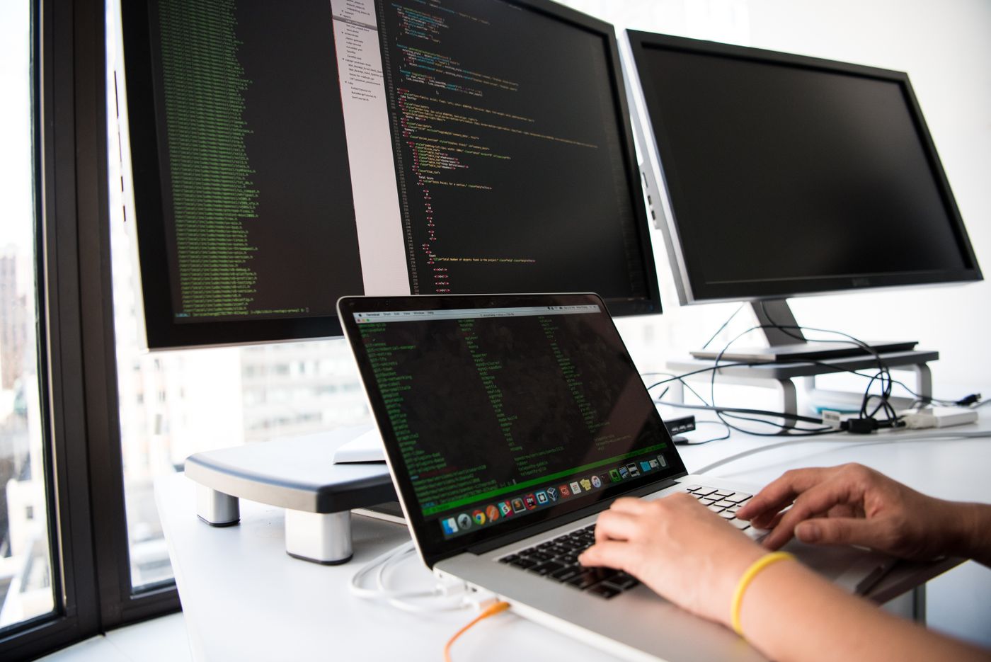 A person typing on a laptop at a desk, with a large monitor behind showing a code editor and a terminal
