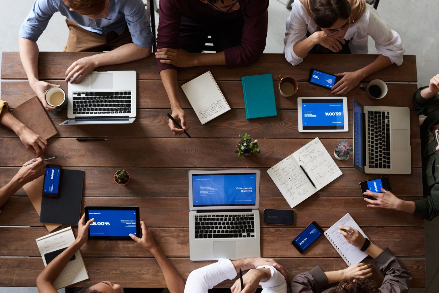 Overhead view of colleagues around a wooden table with laptops, tablets, notebooks and coffee cups during a working session