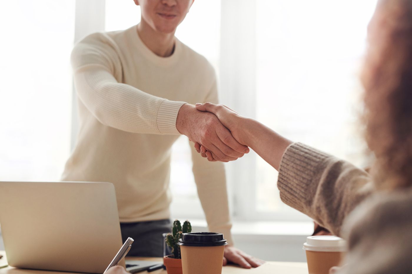 Two people shaking hands across a desk beside a laptop and takeaway coffee cups in a bright office